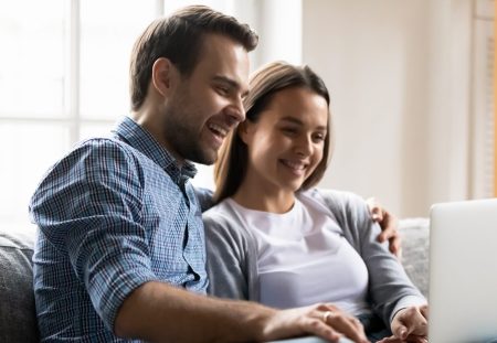 Happy young couple having fun with laptop together close up, sitting on cozy couch at home, looking at screen, chatting or shopping online, laughing man and woman watching movie, enjoying weekend
